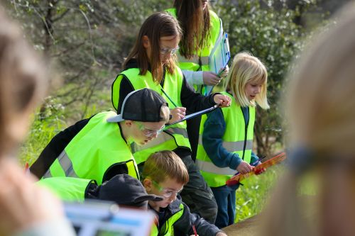 Group of children wearing safety vests and glasses are engaged in an outdoor activity, with some writing on clipboards and others observing.